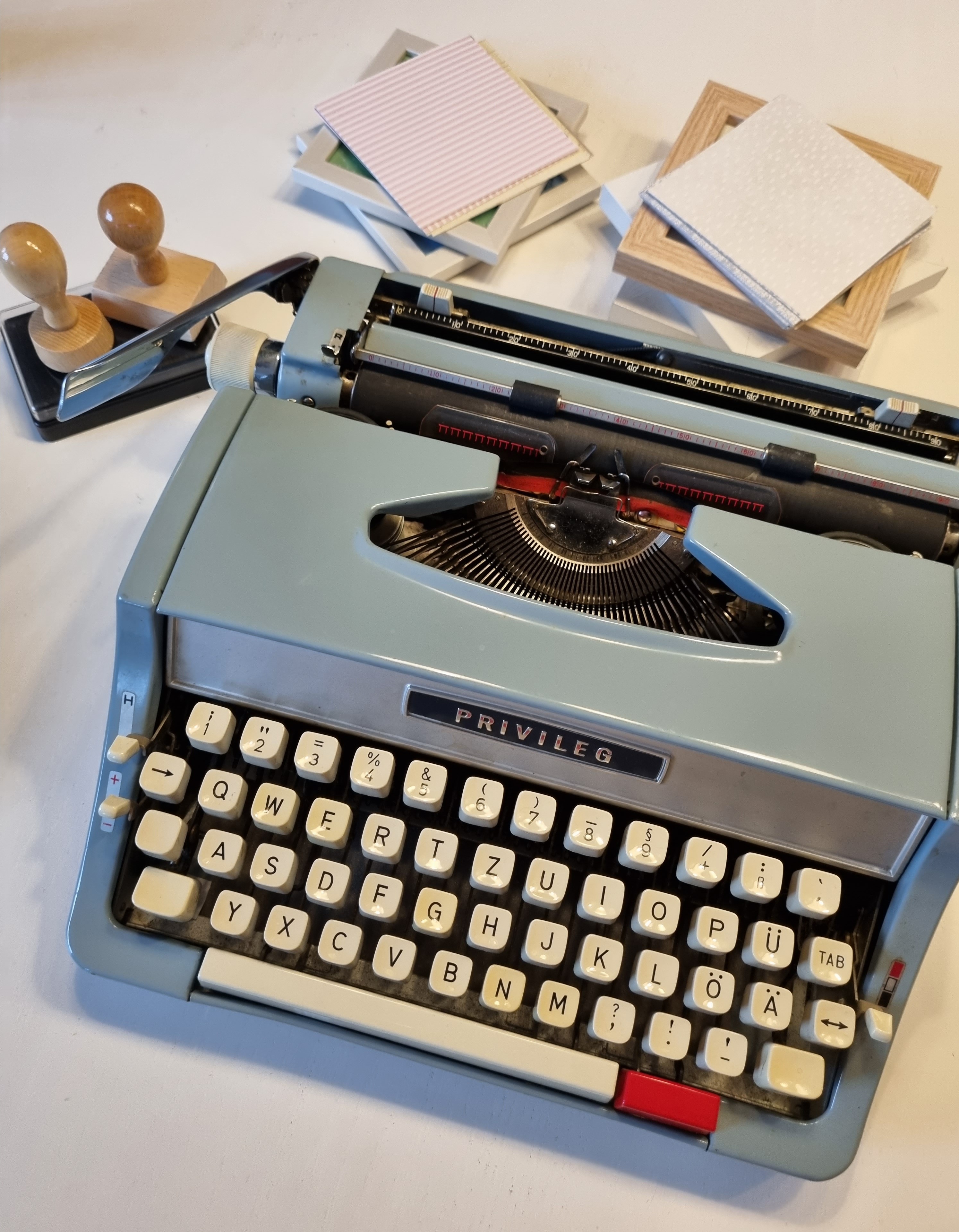 Hellblaue Schreibmaschine „Privileg“ mit Papierunterlagen und zwei Holzstempeln auf einem weißen Tisch arrangiert.