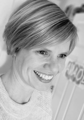 Porträt einer lächelnden Frau in Schwarzweiß, mit kurzem Haar und Strickpullover, im Innenraum vor unscharfem Hintergrund.