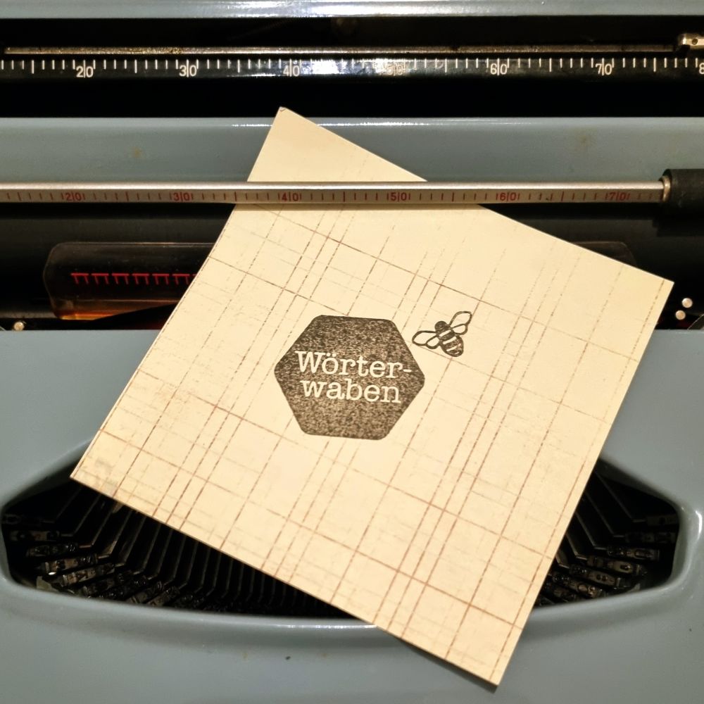 Papier mit „Wörterwaben“-Logo liegt auf einer hellblauen Schreibmaschine mit sichtbarer Skala und Farbband.